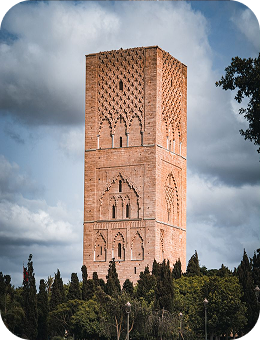 Tall red brick tower with intricate designs above green trees under a cloudy sky.
