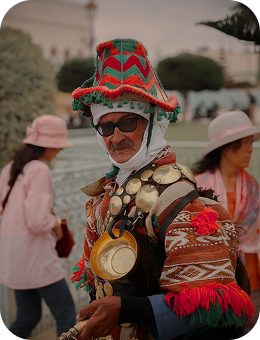 Man in traditional colorful attire and hat with metal ornaments and a brass cup outdoors.