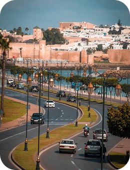 Curved road with cars and motorcycles passing by historic brown city walls under clear blue sky.